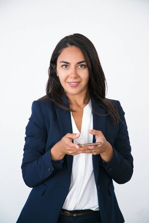 Friendly successful beautiful woman texting message on smartphone. Happy young Latin woman in office jacket using mobile phone and smiling at camera. Communication conceptの写真素材