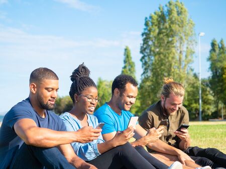 Cheerful friends using smartphones in park. Happy young multiethnic friends sitting in green grass and using mobile phones. Technology conceptの写真素材