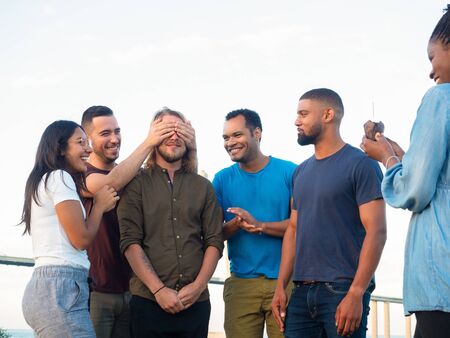 Front view of smiling friends making surprise for young man. Smiling young man covering eyes of his...
