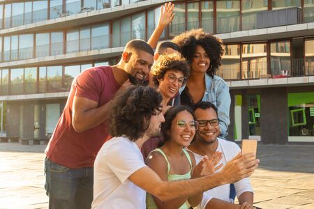 Cheerful long haired guy taking group selfie with his friends. Multiethnic team of friends posing and laughing at phone camera. Fun conceptの写真素材