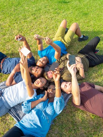 Happy friends lying on grass with smartphones. High angle view of cheerful young multiethnic friends lying on green meadow and taking selfie with cell phones in park. Technology conceptの写真素材
