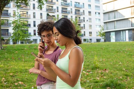 Two female friends walking outside. Women using smartphone and talking on mobile phone, grass and...