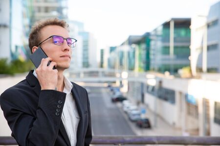 Proud confident businessman calling on cell outside. Young man in office jacket and glasses standing in city street and talking on mobile phone. Phone talk conceptの写真素材
