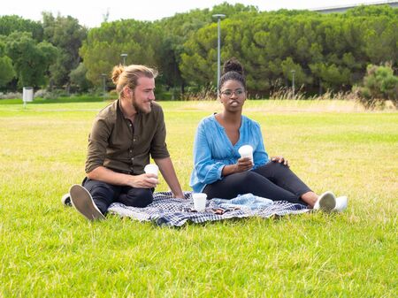 Multiethnic friends resting on lawn in park. Young man and woman holding paper cups and talking while sitting on plaid in park. Weekend conceptの写真素材