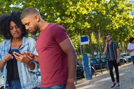 Mix raced couple standing outside and looking at phone screen. Young Caucasian man talking on cell behind them. Communication conceptの写真素材