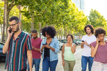 Interracial group of people with smartphone walking outdoors. Men and women going down street, using...