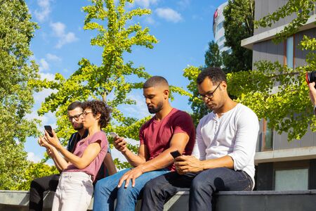 Team of friends discussing their common problems and consulting internet. Men and woman sitting on parapet outdoors and using smartphones. Teamwork conceptの写真素材