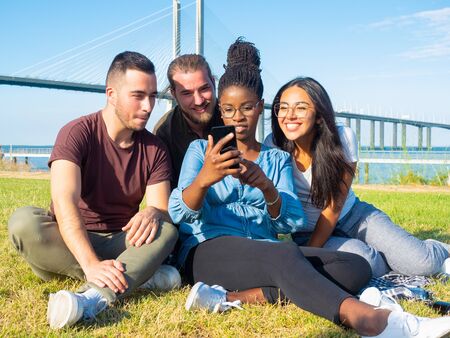 Focused young friends using smartphone outdoor. Smiling multiethnic people sitting on green grass...