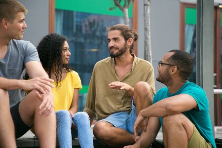 Joyful hipster guy sharing news with his friends outside. Mix raced group of young people sitting on...