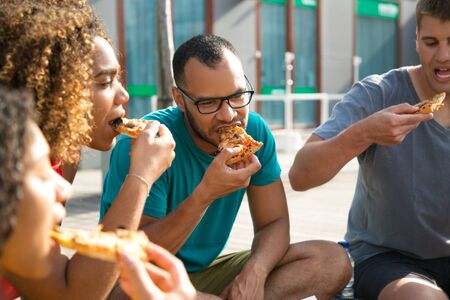 Focused friends excited with pizza eating. Mix raced group of young people sitting outdoors and biting slices. Hungry urban people conceptの写真素材