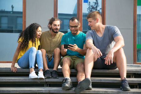 Excited friends watching video on mobile phone. Cheerful Latin guy using smartphone and showing...