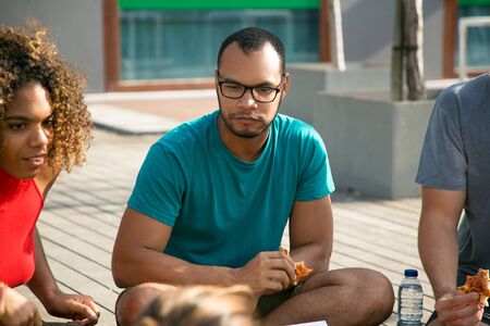 Pensive Latin guy enjoying pizza in friendly company. Group of young people sitting outdoors and...
