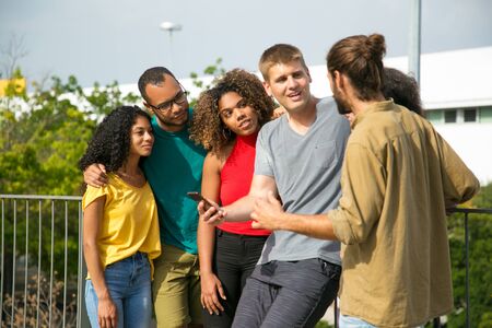 Excited Caucasian guy with smartphone arguing with male friend. Mix raced team of people standing outdoors, leaning on railing, talking, listening, arguing. Using phone conceptの写真素材