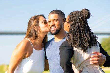 Two female friends kissing African American man on meadow. Three friends spending time together in...