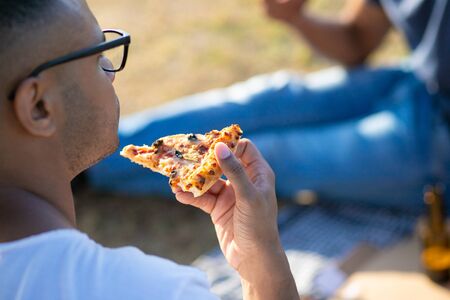 Side view of young man biting pizza. Young guy in eyeglasses having picnic with friends. Concept of picnicの写真素材