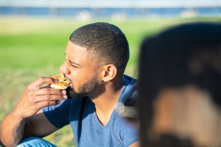 Handsome bearded man biting piece of pizza. African American guy sitting on meadow and eating pizza. Concept of picnicの写真素材