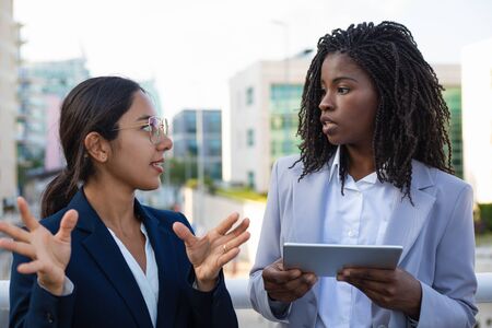 Coworkers talking and using tablet pc. Young multiethnic businesswomen holding digital tablet and looking at each other outdoor. Technology conceptの写真素材