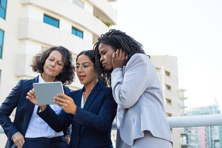 Focused female colleague with tablet pc. Low angle view of professional multiethnic businesswomen standing together on street and using tablet computer. Technology conceptの写真素材