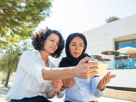 Positive female coworkers taking selfie outside. Multicultural business women using smartphone for video call. Communication conceptの写真素材