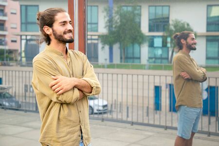 Happy hipster guy waiting someone outside. Young man in casual with bun and stubble standing in city...
