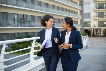 Smiling businesswomen with papers outdoor. Cheerful multiethnic female colleagues holding documents, talking and walking on street. Cooperation conceptの写真素材
