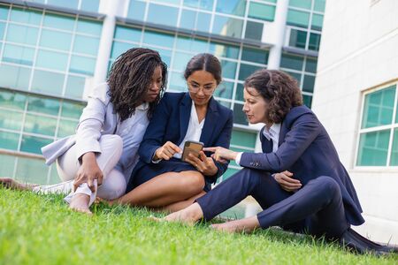 Focused businesswomen using smartphone outdoor. Multiethnic barefoot female colleagues sitting on green lawn and using mobile phone. Technology conceptの写真素材