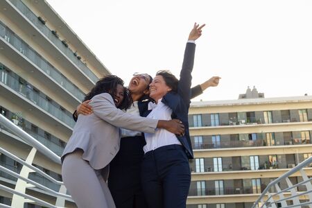 Happy businesswomen hugging outdoor. Low angle view of cheerful multiethnic female colleagues triumphing and hugging on street. Success conceptの写真素材