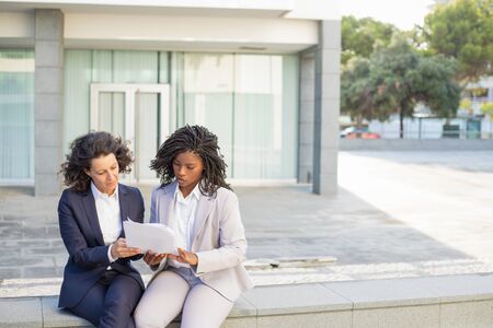 Professional businesswomen working with papers. Serious multiethnic female colleagues sitting together and discussing documents outdoor. Paperwork conceptの写真素材