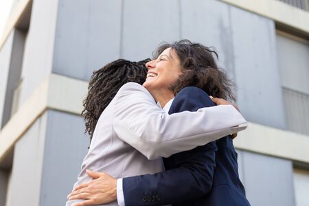 Two smiling colleagues hugging on street. Cheerful female friends meeting during break. Communication conceptの写真素材