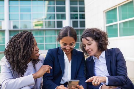 Happy businesswomen with smartphone outdoor. Cheerful multiethnic female colleagues sitting together and using cell phone outdoor. Technology conceptの写真素材