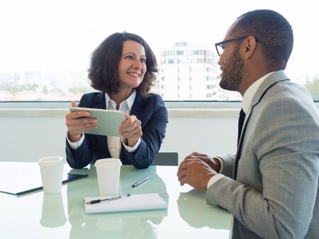 Two excited coworkers discussing team project in meeting room. Happy business woman holding tablet...