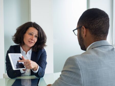 Two business people watching content on tablet at meeting table. Business woman sitting against male colleague and showing tablet screen. Wireless technology conceptの写真素材