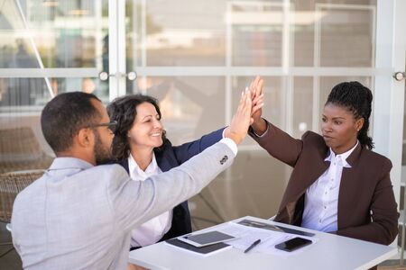 Happy businesspeople celebrating team success. Multiethnic group of man and women sitting at table...