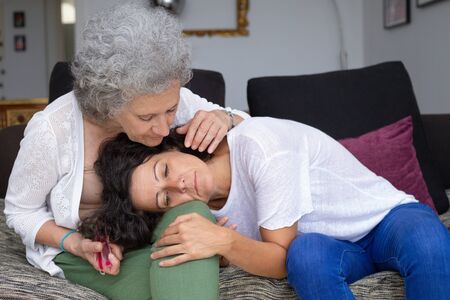 Old mother supporting daughter. Senior mother sitting on couch and looking down at unhappy adult...