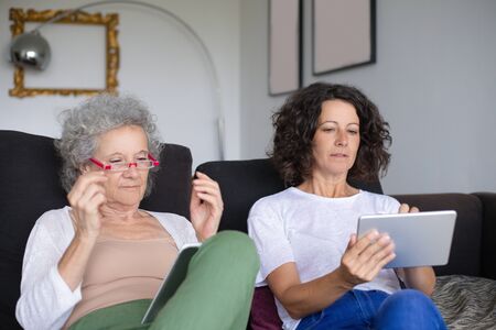 Focused mother and daughter with digital tablets. Senior and middle aged women sitting on couch and using tablet computers at home. TEchnology conceptの写真素材