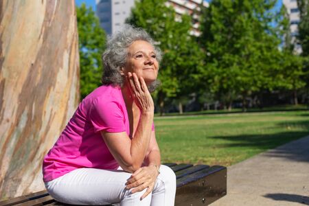 Joyful old lady waiting someone in city park. Senior grey haired woman in casual sitting on bench outdoors, leaning chin on hand and looking into distance. City park conceptの写真素材
