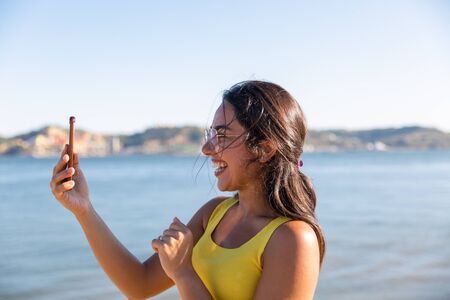 Happy joyful woman enjoying video call outside. Beautiful young Latin woman standing near river or bay, holding mobile phone, speaking at screen and laughing. Video call conceptの写真素材