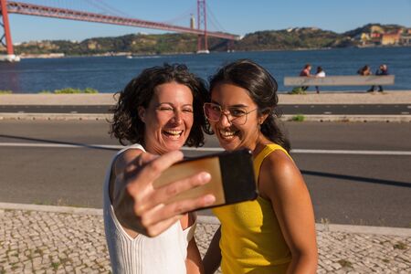 Happy excited female friends posing for smartphone camera outdoors. Two women standing on promenade,...