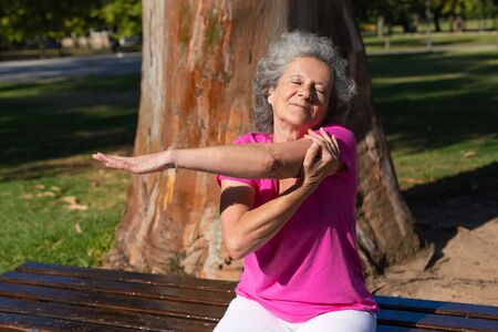 Serene old lady warming up for morning exercise in park. Senior grey haired woman in casual sitting on bench and stretching hand. Age and activity conceptの写真素材
