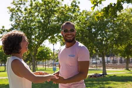 Happy couple meeting in park. Joyful man and woman standing outdoors against each other, holding hands and laughing. Lovely meeting conceptの写真素材