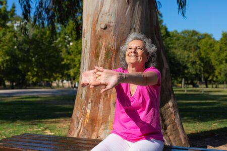 Happy old lady doing morning exercise outdoors. Senior grey haired woman in casual sitting on bench and stretching arms. Fitness at senior age conceptの写真素材