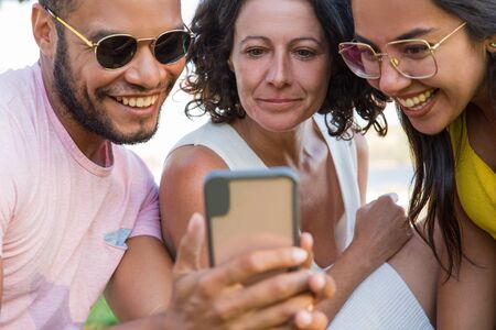Team of cheerful friends watching content on cellphone together. Man and women sitting outdoors and looking at phone screen. Media content conceptの写真素材
