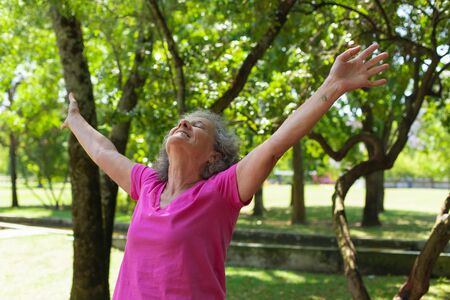 Happy carefree old lady exercising outdoors. Senior grey haired woman in casual with closed eyes standing in park, raising hands and looking up. Leisure outdoors conceptの写真素材