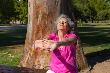 Peaceful old lady enjoying outdoor workout in park. Senior grey haired woman in casual sitting on bench and stretching arms. Workout in park conceptの写真素材