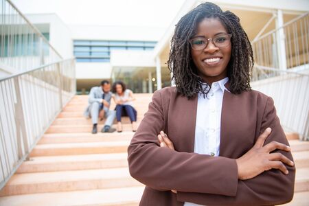 Cheerful African American businesswoman. Portrait of happy young businesswoman standing with crossed arms and smiling at camera while colleagues sitting behind on steps. Business conceptの写真素材