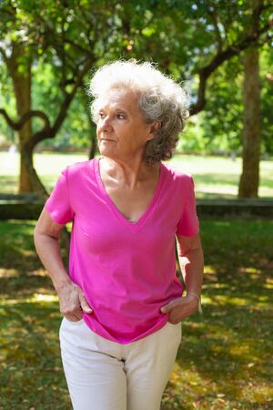 Pensive old lady enjoying leisure time in park. Senior grey haired woman in casual walking on grass and looking away. Age and active life style conceptの写真素材