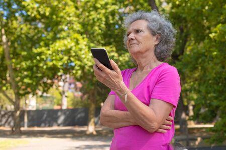 Serious focused old lady listening to voice message on phone in park. Senior grey haired woman in...