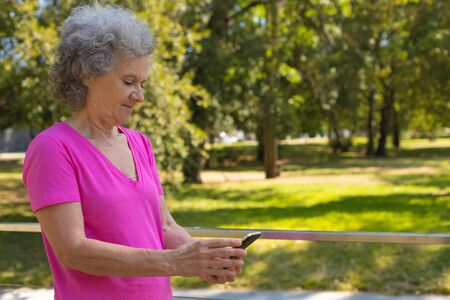 Positive focused old lady texting message while walking on parkway. Happy senior grey haired woman in casual using mobile phone in park. Wireless connection conceptの写真素材