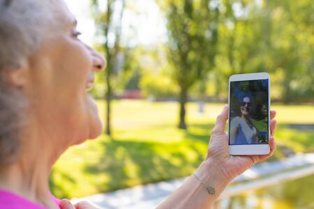 Cheerful old lady chatting with daughter through video call while walking in park. Senior grey haired woman in casual using mobile phone with female face on screen. Video call conceptの写真素材