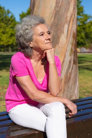 Pensive tired old lady waiting someone in park. Senior grey haired woman in casual sitting on bench, leaning head on hand and looking away. Waiting in park conceptの写真素材